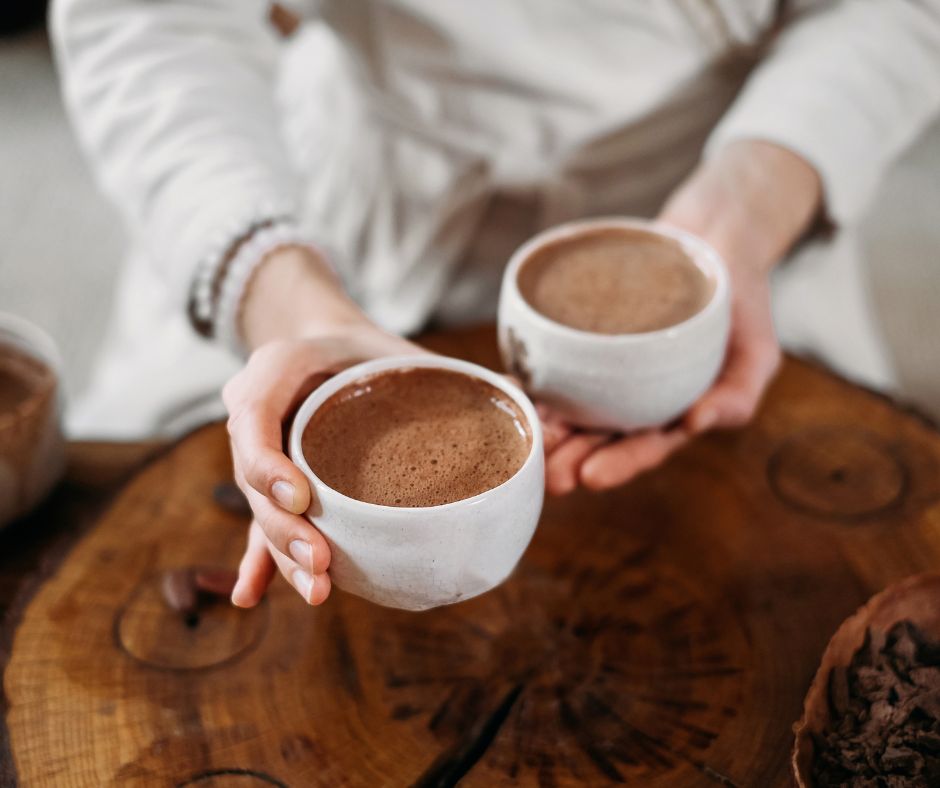 Auf dem Bild sind im Vordergrund zwei Frauenhände zu sehen. Sie hält in der rechten und der linken Hand jeweils eine Tasse Kaffee oder Kakao. Die eine Hand ist etwas weiter vorne im Bild, so als würde sie die Tasse ihrem Gegenüber anbieten.